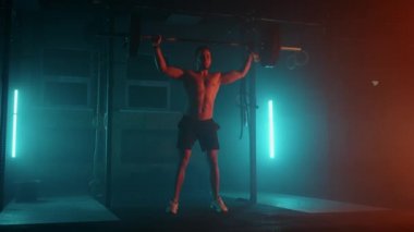 Mavi-turuncu neon ışıklı erkek kaldırıcı spor salonunda antrenmana hazırlanıyor. Çıplak gövdeli güçlü bir adam halteri başının üzerine kaldırır. Ağırlık kaldırma egzersizi yapar..