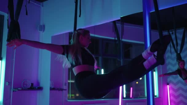 Une femme fait du yoga sportif dans l'air sur un hamac dans la lumière au néon effectuant des exercices gracieux et des mouvements d'étirement et de danse au ralenti. — Video