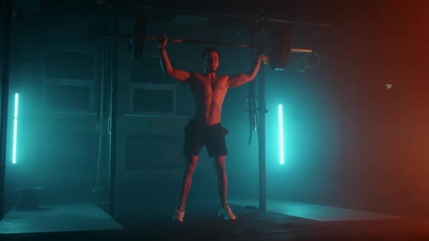 Homme powerlifter dans une lumière néon bleu-orange se prépare pour une séance d'entraînement dans la salle de gym. Un homme fort avec un torse nu fait soulever une haltère au-dessus de sa tête, un exercice de secousse de l'haltérophilie. — Video