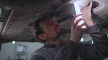 Otomobil tamircisi yavaş çekimde arıza aramak için asansörde kaldırılan arabanın altına bakar.