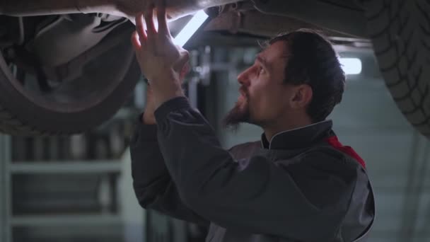 Człowiek auto mechanik patrzy pod samochód podniesiony na dźwigu w poszukiwaniu usterek w zwolnionym tempie — Wideo stockowe