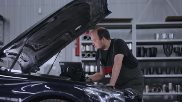 Ein Automechaniker auf der Motorhaube eines Autos mit einem Tablet diagnostiziert alle Systeme und sucht nach Störungen — Stockvideo