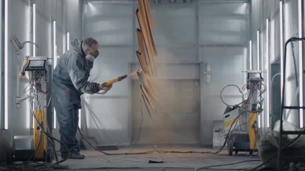 Een man in een beschermend pak en masker spuit verf door een spuitpistool in slow motion op stalen onderdelen. — Stockvideo