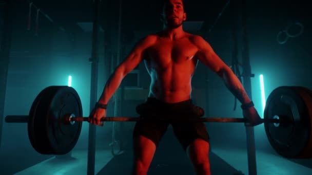 Męski podnośnik w niebiesko-pomarańczowym świetle neonowym przygotowuje się do treningu w siłowni. Silny mężczyzna z nagim tułowiem podnosi sztangę nad głowę, ćwiczy szarpanie od podnoszenia ciężarów.. — Wideo stockowe