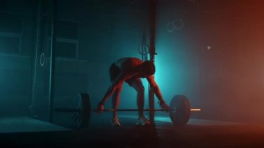 Mavi-turuncu neon ışıklı erkek kaldırıcı spor salonunda antrenmana hazırlanıyor. Çıplak gövdeli güçlü bir adam halteri başının üzerine kaldırır. Ağırlık kaldırma egzersizi yapar..