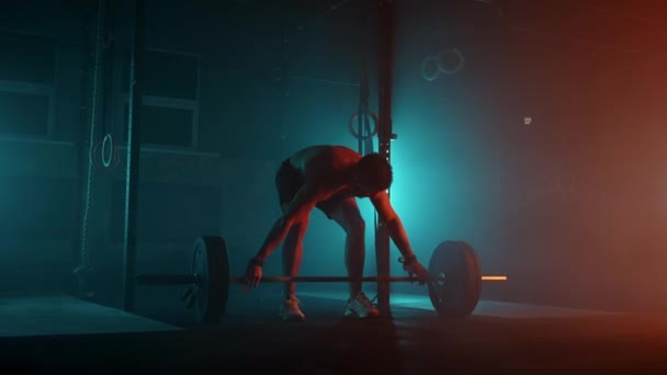 El levantador de pesas masculino en una luz de neón azul-naranja se está preparando para un entrenamiento en el gimnasio. Un hombre fuerte con un torso desnudo levanta una barra sobre su cabeza, un ejercicio de tirón de levantamiento de pesas. — Vídeo de stock