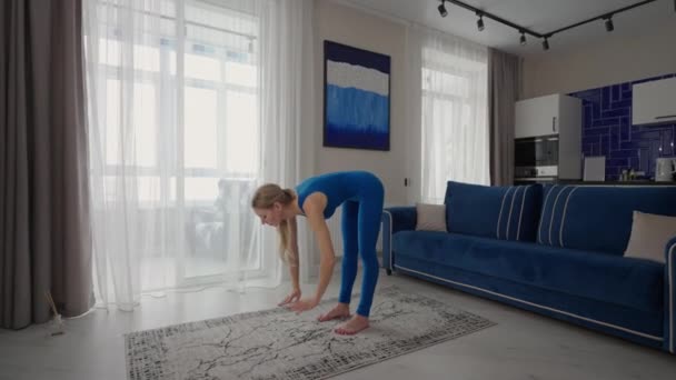 Una mujer en casa hace ejercicios sin salir de casa. Entrenamiento en casa. Una mujer entrena en la alfombra con un traje azul — Vídeo de stock