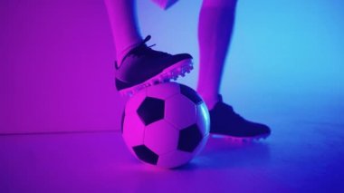 Stüdyonun mavi-kırmızı neon ışığı altında ağır çekimde topun üzerinde duran profesyonel bir siyahi futbolcunun ayağına yakın çekim. Brezilyalı futbolcu poz vermek için topa bastı
