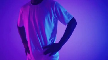 Beyaz üniformalı siyahi bir futbolcunun yavaş çekim portresi stüdyonun mavi-kırmızı neon ışığı altında kameraya bakıyor. Kendine güvenen ve muzaffer acımasız bir futbolcu.