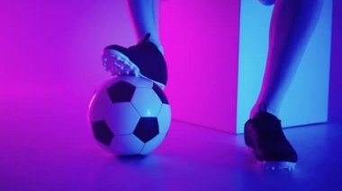 Stüdyonun mavi-kırmızı neon ışığı altında ağır çekimde topun üzerinde duran profesyonel bir siyahi futbolcunun ayağına yakın çekim. Brezilyalı futbolcu poz vermek için topa bastı