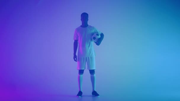 Retrato en cámara lenta de un jugador de fútbol profesional negro con un uniforme blanco mirando a la cámara en la luz de neón azul-rojo del estudio. Un jugador de fútbol brutal confiado y victorioso — Vídeos de Stock