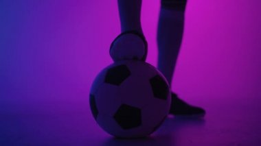 Stüdyonun mavi-kırmızı neon ışığı altında ağır çekimde topun üzerinde duran profesyonel bir siyahi futbolcunun ayağına yakın çekim. Brezilyalı futbolcu poz vermek için topa bastı