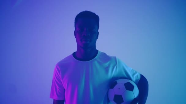 흰색 유니폼을 입은 흑인 프로 축구 선수의 느린 움직임 사진 이 스튜디오의 파란색 네온 조명 속에서 카메라를 들여다보고 있습니다. 자신감 있고 승리 한 잔인 한 축구 선수 — 비디오