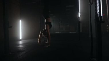 Güçlü genç bir atletik kadın spor salonunda baş aşağı durur. Bir kadın karanlık bir spor salonunda eli ayağına dolaşır. En azından baş aşağı çalışıyorum.