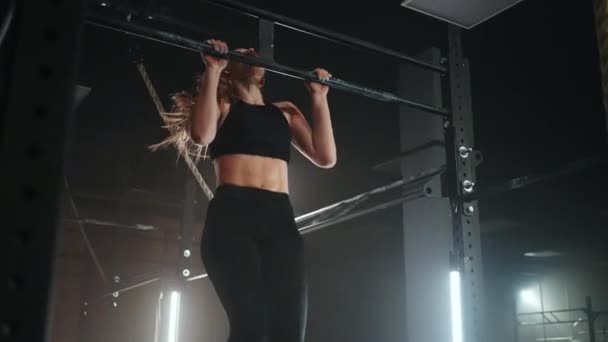 A sporty young woman pulls herself up on a horizontal bar in a dark gym in a beautiful neon backlight. Endurance and perseverance in pulling up movement towards the goal — Stock Video