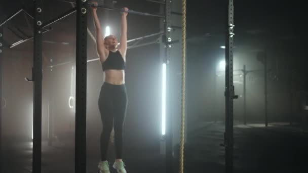 Una joven deportista se acerca a una barra horizontal en un gimnasio oscuro con una hermosa luz de fondo de neón. Resistencia y perseverancia en levantar el movimiento hacia la meta — Vídeo de stock
