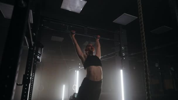 En sportig ung kvinna drar sig upp på en horisontell bar i ett mörkt gym i ett vackert neonljus. uthållighet och uthållighet i att dra upp rörelsen mot målet — Stockvideo