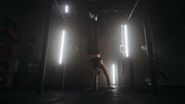 Silna młoda atletyczna kobieta idzie do góry nogami na siłowni. Kobieta jest zdrowa na rękach w ciemnej siłowni. Szkolenie co najmniej do góry nogami — Wideo stockowe