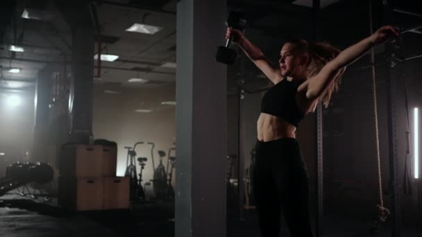 В замедленной съемке женщина поднимает гантель над головой во время тренировки в тёмном спортзале. Спортивная сильная женщина выполняет сложную тренировку с поднятием гантели — стоковое видео