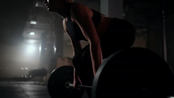Zeitlupe: Eine Frau hebt in einem dunklen Fitnessstudio eine Langhantel mit viel Gewicht. Eine starke junge Frau stemmt Gewichte für ein Workout — Stockvideo