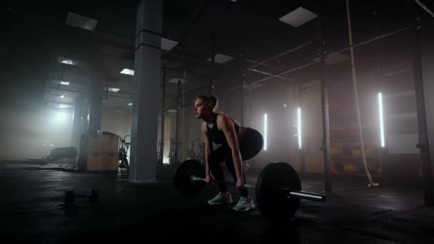 Zeitlupe: Eine Frau hebt in einem dunklen Fitnessstudio eine Langhantel mit viel Gewicht. Eine starke junge Frau stemmt Gewichte für ein Workout — Stockvideo