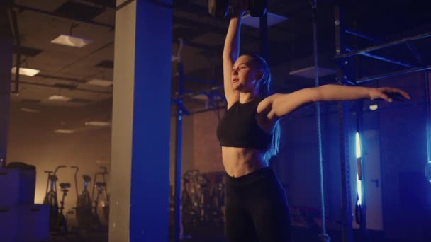 In slow motion, a woman lifts a dumbbell over her head while working out in a dark gym. Athletic strong woman performs a difficult workout with lifting dumbbells — Stock Video