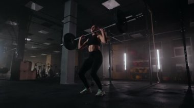 Ağır çekimde, bir kadın koyu renk bir spor salonunda halter kaldırıyor. Güçlü genç bir kadın egzersiz yapmak için ağırlık kaldırır.