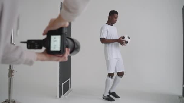 Sesión de fotos entre bastidores: Un fotógrafo en un estudio fotográfico utiliza un flash para fotos de un jugador de fútbol profesional negro. Sesión de fotos en el estudio de una revista deportiva, publicidad — Vídeo de stock