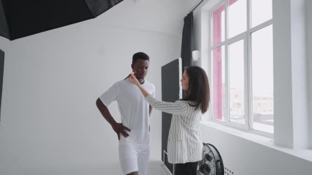 Detrás de las escenas de una sesión de fotos: Un hermoso jugador de fútbol modelo negro se está preparando para una sesión de fotos para una revista, un artista de maquillaje corrige el tono de la piel para las fotos en una revista deportiva — Vídeo de stock