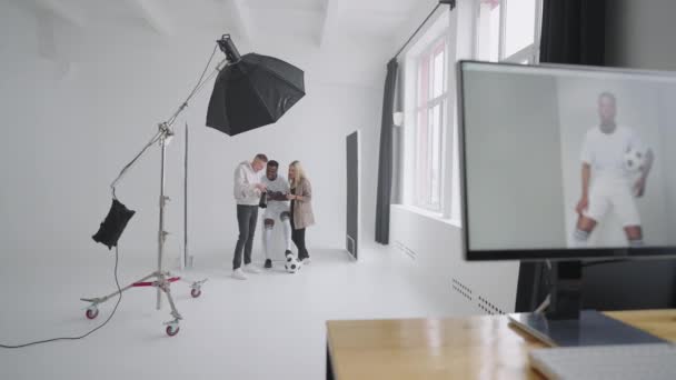 Οθόνη με διαφημιστικό εξώφυλλο της φωτογραφίας Στο φόντο ένας φωτογράφος, ένας ποδοσφαιριστής, ένα μοντέλο και ένας σκηνοθέτης κοιτούν φωτογραφίες από μια φωτογράφιση για το εξώφυλλο μιας κάμερας — Αρχείο Βίντεο