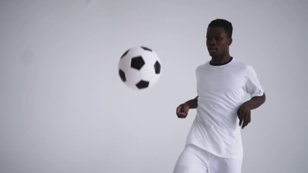 白い背景に白いユニフォームを着たプロの黒人サッカー選手がスローモーションでボールをジャグリングする。サッカーボールを持つアフリカ系アメリカ人の民族のサッカー選手 — ストック動画