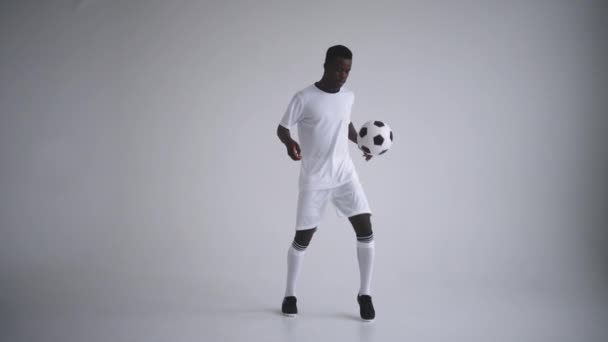 Um jogador profissional de futebol preto em um uniforme branco em um fundo branco faz malabarismos com uma bola em câmera lenta. Jogador de futebol étnico afro-americano com bola de futebol — Vídeo de Stock