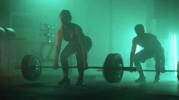 A man and a woman in a colored neon red and blue light in slow motion lift heavy barbells over their heads. Strength and power training with heavy weight. The concept of athletic and strong couples — Stock Video