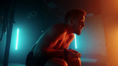 Egzersiz sonrası yorgun bir adam halterde oturur ve ağır çekimde mavi-turuncu renklerle spor salonunda dinlenir.