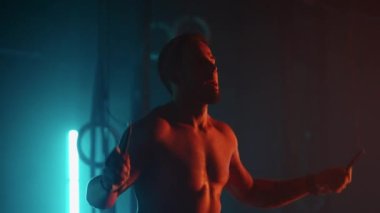 Tişörtsüz, çıplak gövdeli kaslı bir adam ağır çekimde ipe atlıyor. Bir dövüşçü ipin üzerinden atlar.