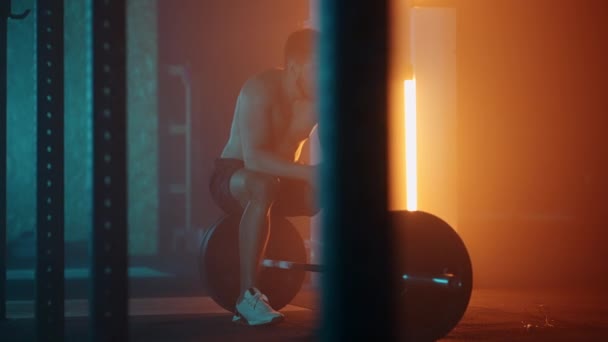 En trött man efter träning sitter på en skivstång och vilar i gymmet i blå-orange färger i slow motion — Stockvideo