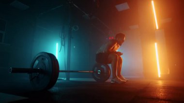 Egzersiz sonrası yorgun bir adam halterde oturur ve ağır çekimde mavi-turuncu renklerle spor salonunda dinlenir.