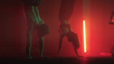 Bir erkek ve bir kadın spor salonunda ağır çekimde amuda kalkarlar. Güçlü bir adam ve amuda kalkmış bir kadın spor salonunun zemininde renkli gaz lambalarıyla yürüyorlar.