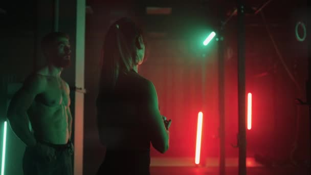 Dva muži a žena v tělocvičně dělají zatáčky na vodorovném baru společně v tmavém neonovém světle. Atmosféra fitness a školení — Stock video