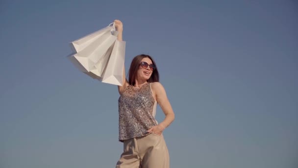 Shopaholic γυναίκα κρατά τσάντες ψώνια ενάντια στον γαλάζιο ουρανό — Αρχείο Βίντεο