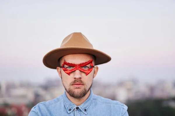 Poważny superbohater mężczyzna w czerwonych oczu maska i kapelusz z widokiem na miasto — Zdjęcie stockowe
