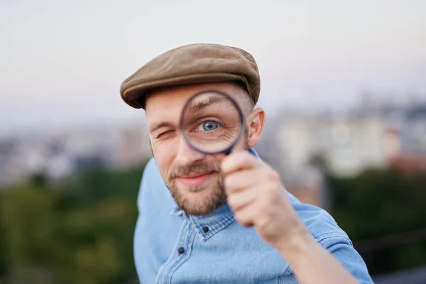 Détective mâle dans le chapeau de pointe faisant grand oeil en utilisant la loupe Image En Vente