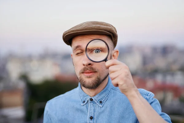 Man in peaked cap looking at camera using magnifying glass — Stock Photo, Image