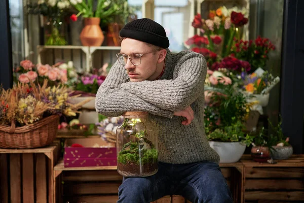 Stylish caucasian tired entrepreneur florist male wearing sweater, beanie and eyeglasses in flower shop. Handsome greenhouse owner or gardener with flowers and bouquets in shop. High quality image
