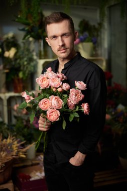 Stylish caucasian macho male with mustache holding pink roses bouquet in hand, looking at camera wearing black shirt in flower store with dark background. Romantic or dating concept. Vertical photo