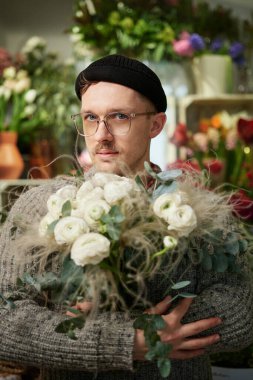 Good looking male florist wearing sweater, beanie and eyeglasses holding flower bouquet. Small business concept. Bearded man with bouquet of white roses in flower shop. High quality vertical image
