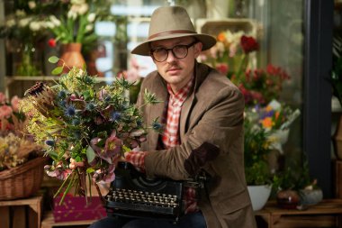 Cute male person in eyeglasses and hat sitting with old typewriter in flower shop. Poet or writer working concept. High quality vertical image