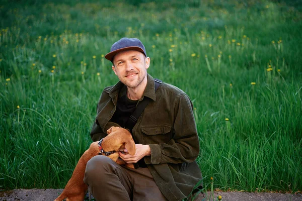 Cute male sitting and playing with small brown puppy in park with green grass. Pets as family members concept image. Handsome young man with dachshund outdoors. Man on a green grass with dog
