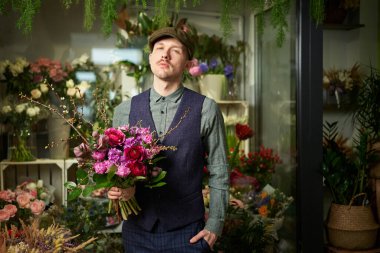 Serious emotion caucasian male in peaked cap and vintage clothes from 20s holding beautiful bouquet of red and violet flowers. Congratulating with flowers. Mothers Day or Valentines Day concept