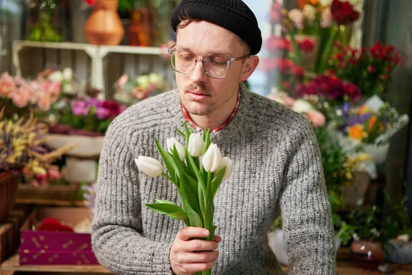 Portrait of attractive bearded male in gray sweater, eyeglasses and beanie hat holding beautiful bouquet of spring white tulips. Mothers Day, Valentines or International Womens Day concept